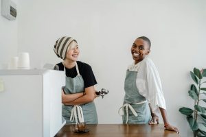 Two people in aprons stand behind a counter. One holds a portafilter, and both are smiling. A coffee machine is on the counter, with a potted plant visible in the background.