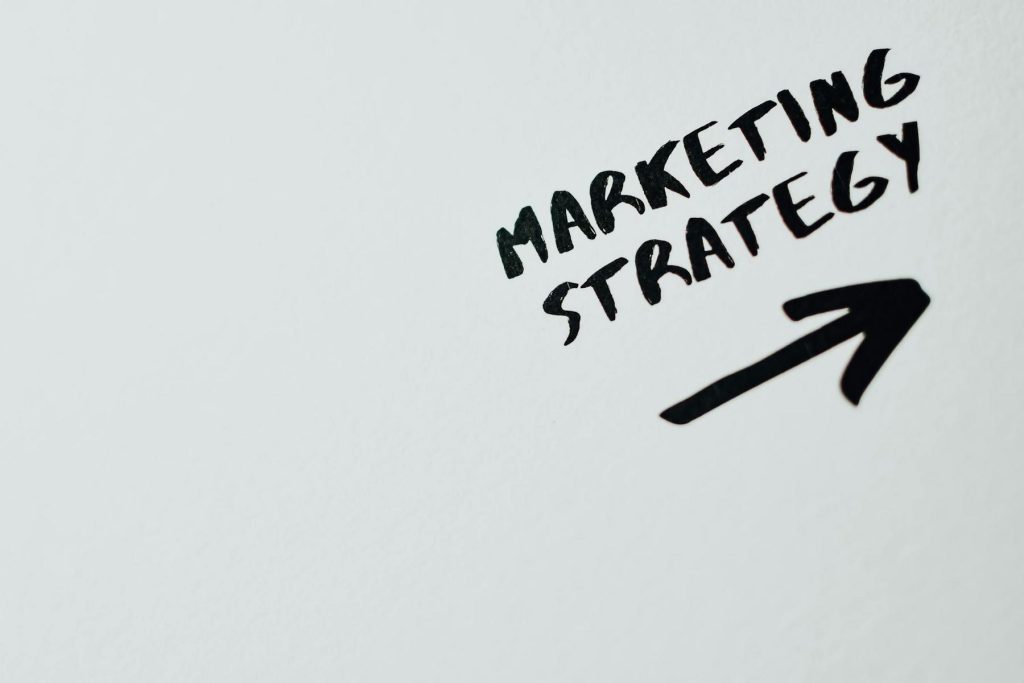 The image shows the words "MARKETING STRATEGY" handwritten in black on a white surface with an arrow pointing to the right.