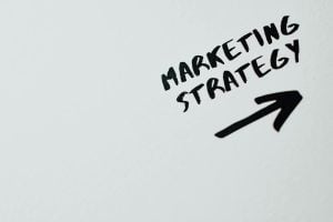 The image shows the words "MARKETING STRATEGY" handwritten in black on a white surface with an arrow pointing to the right.