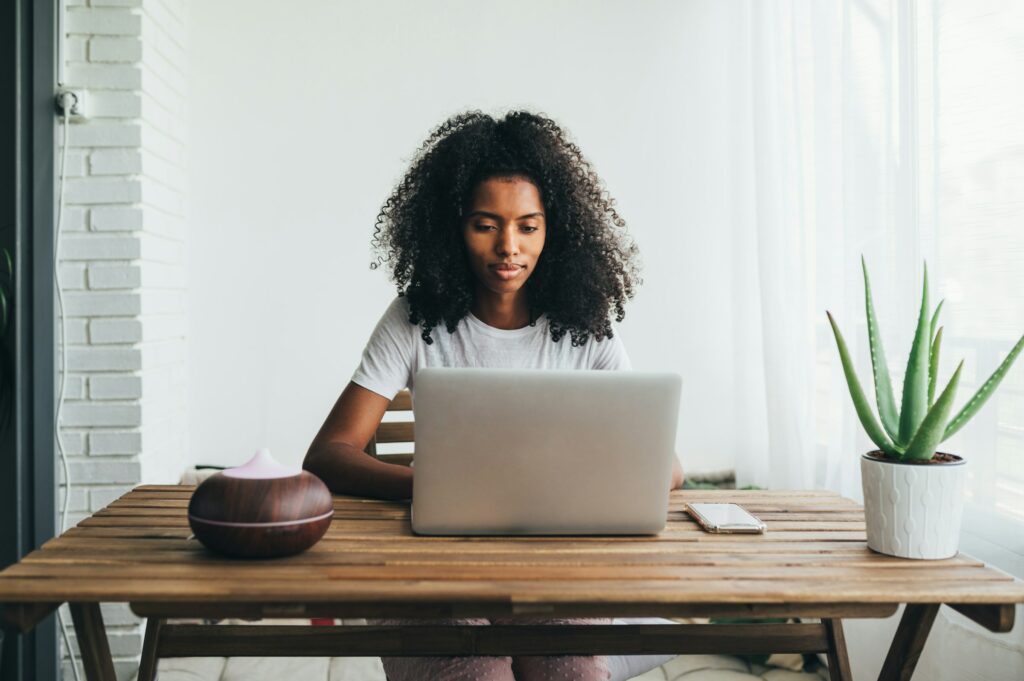 A Young Woman Sitting At A Table Working On Her Laptop, Utilizing Home Office Essentials.