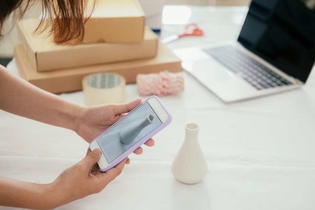 A woman using a cell phone in front of a laptop to sell products.