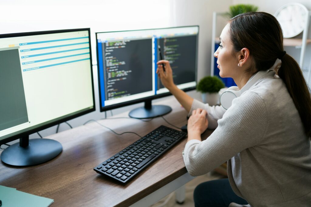 A woman is working on a computer with two monitors, coding in CSS.