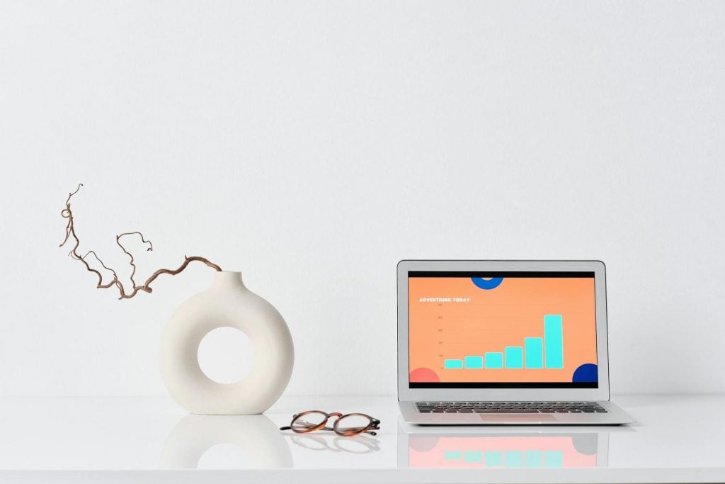 Laptop displaying a bar graph on a desk with a modern vase and eyeglasses, set against a plain white background.