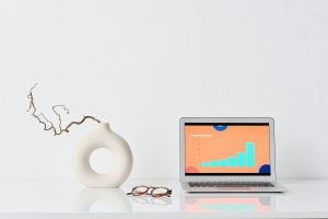 Laptop displaying a bar graph on a desk with a modern vase and eyeglasses, set against a plain white background.