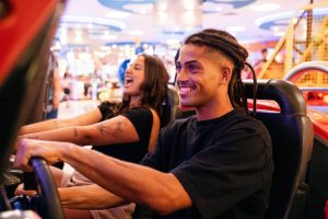 In a lively arcade, two friends immerse themselves in a thrilling car racing game. The man is focused and smiling, while the woman laughs gleefully in the background, capturing the joy of New Zealand's vibrant entertainment scene—a promising future outlook for leisure and fun.