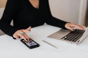 A person uses a smartphone and works on a laptop at a white desk, with a pen nearby.