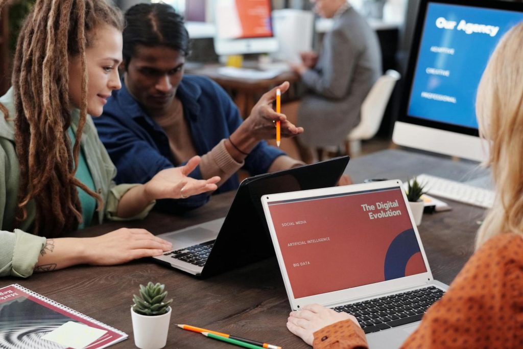 People collaborating at a table with laptops. The screen displays "The Digital Evolution"; a presentation with topics like social media and AI. Small plants and writing materials are on the table.