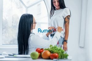 A healthcare professional from Rumen measures the waist of a standing woman in a bright room, where a table adorned with fruits and vegetables signifies an integrated digital presence focused on holistic wellness.