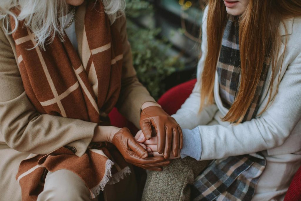 Two people sitting closely, wearing coats and scarves. One person has leather gloves and is holding the other's hand.