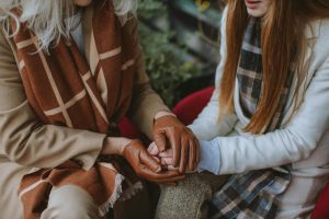 Two people sitting closely, wearing coats and scarves. One person has leather gloves and is holding the other's hand.