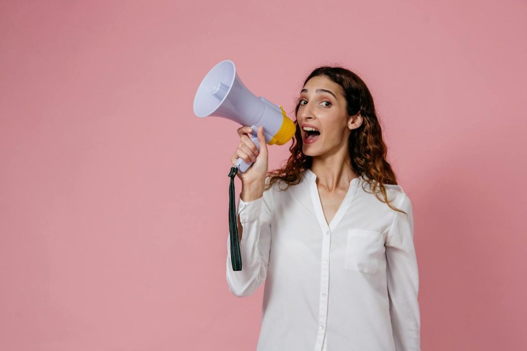 Woman with long hair holds a megaphone and smiles against a pink background, wearing a white button-up shirt.
