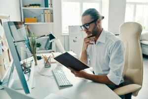 A man in glasses is reading a book in front of a computer, immersed in the enigma of business.