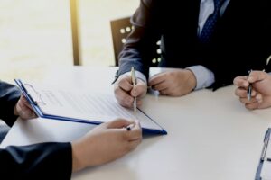 Three people in suits are seated at a table, with one person signing a document on a clipboard labeled "What is SLA" while the others observe and hold pens.